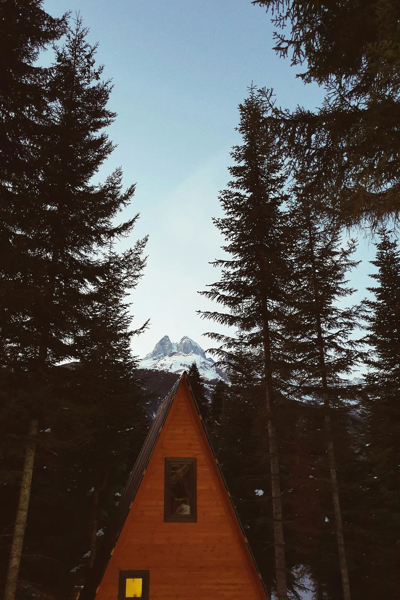 Triangular cabin house framed by pine trees, watched over by Ushba mountain in the background